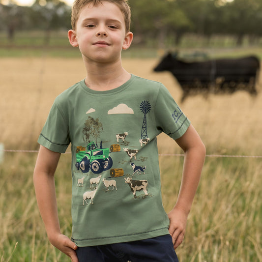 A young boy wearing a sage green graphic t-shirt with a farm-themed print (tractor, cows, sheep, windmill) and navy blue shorts, standing in a grassy field with a blurred cow in the background.
