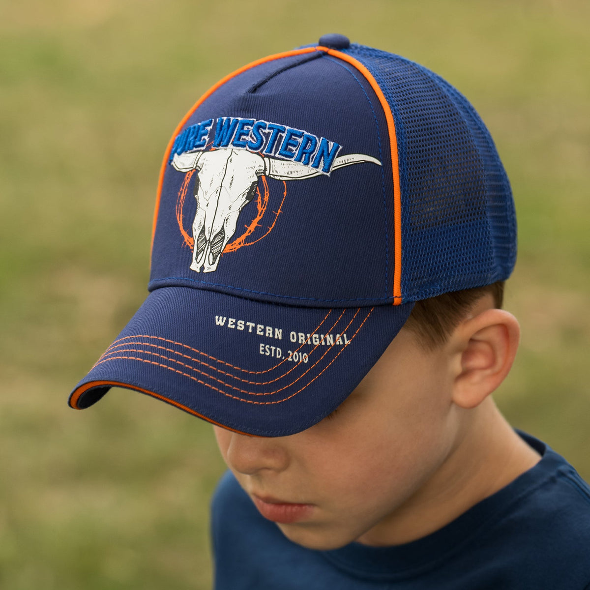 A boy wearing a navy blue trucker hat with orange piping and a white longhorn bull skull logo that reads 'WE WESTERN'.