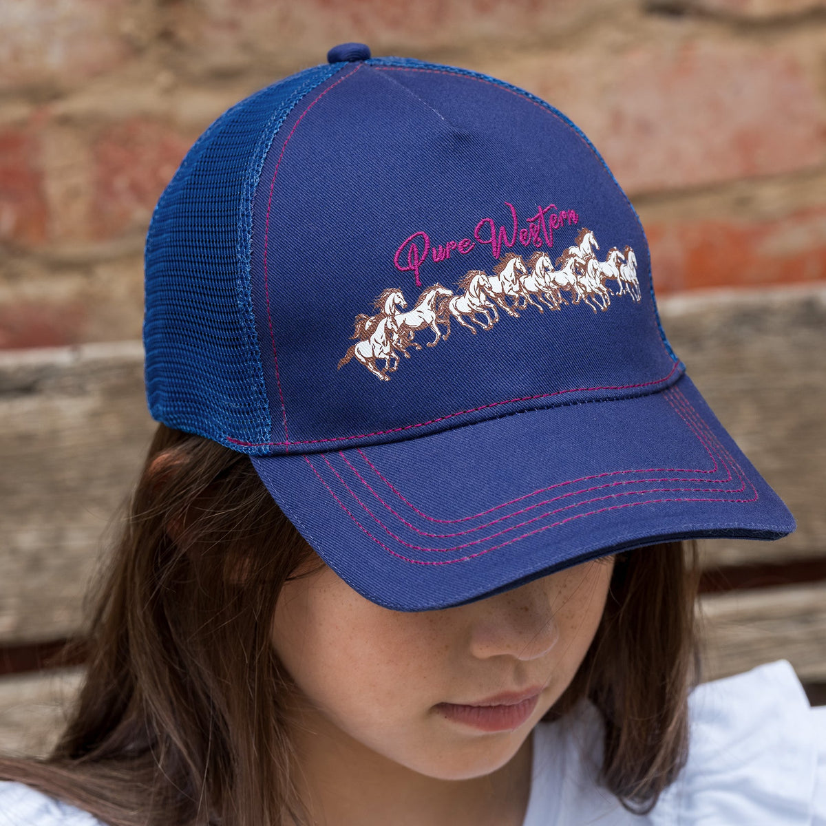 A child wearing a navy blue trucker-style baseball cap with pink and white embroidery that reads 'Pure Western' above a line of white horses. The cap has a navy front panel, pink piping, and a navy mesh back.