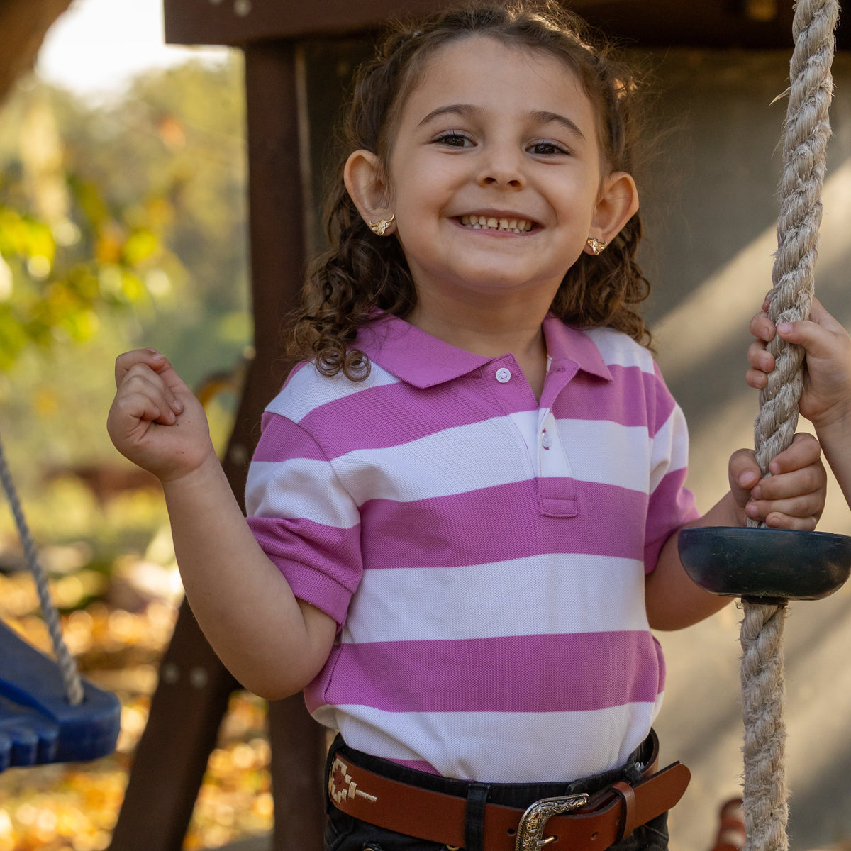 A young girl smiling on an outdoor playground, wearing a pink and white striped polo shirt, dark pants, and a brown belt while holding a rope swing.