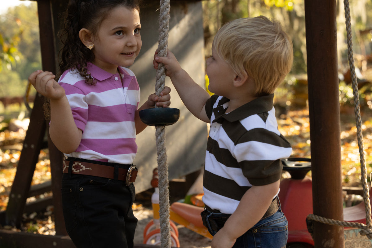 Two young children standing on a playground swing set, wearing matching striped polo shirts. The girl wears a pink and white striped shirt with dark pants, and the boy wears a black and white striped shirt with blue jeans.