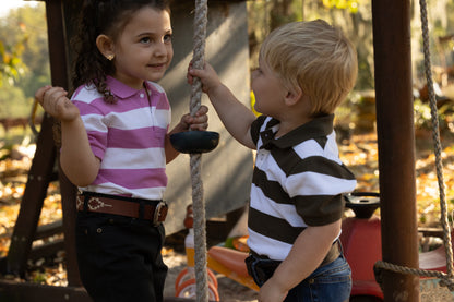 Two young children standing on a playground swing set, wearing matching striped polo shirts. The girl wears a pink and white striped shirt with dark pants, and the boy wears a black and white striped shirt with blue jeans.