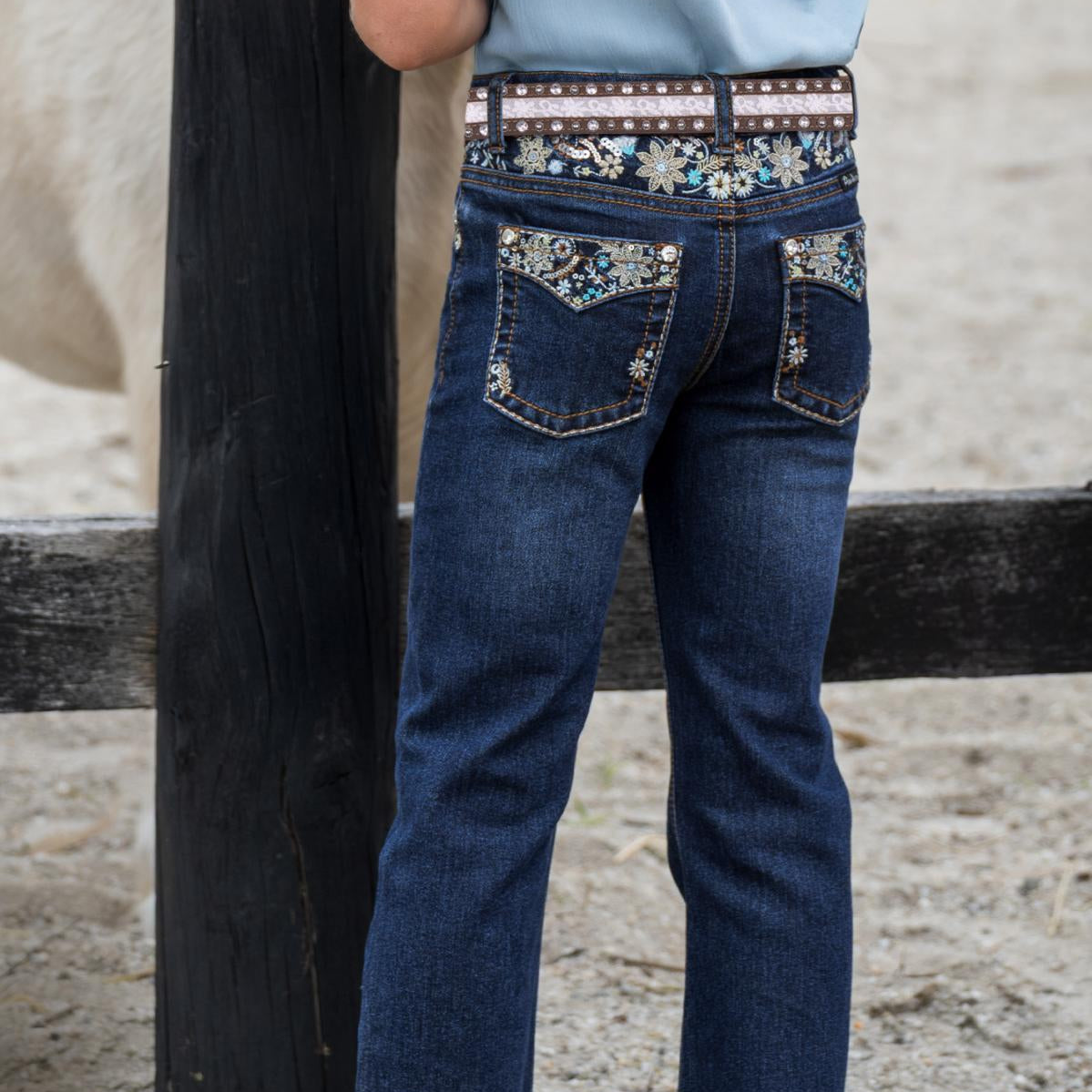 A child wearing dark blue denim jeans with decorative floral embroidery on the back pockets and a patterned belt, standing outdoors next to a wooden fence.