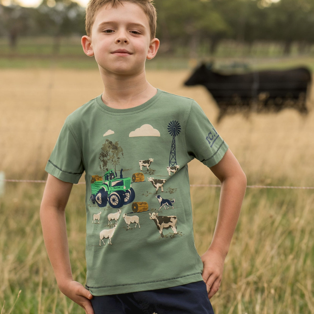 A young boy wearing a sage green graphic t-shirt with a farm-themed print (tractor, cows, sheep, windmill) and navy blue shorts, standing in a grassy field with a blurred cow in the background.