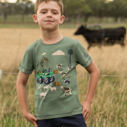 A young boy wearing a sage green graphic t-shirt with a farm-themed print (tractor, cows, sheep, windmill) and navy blue shorts, standing in a grassy field with a blurred cow in the background.