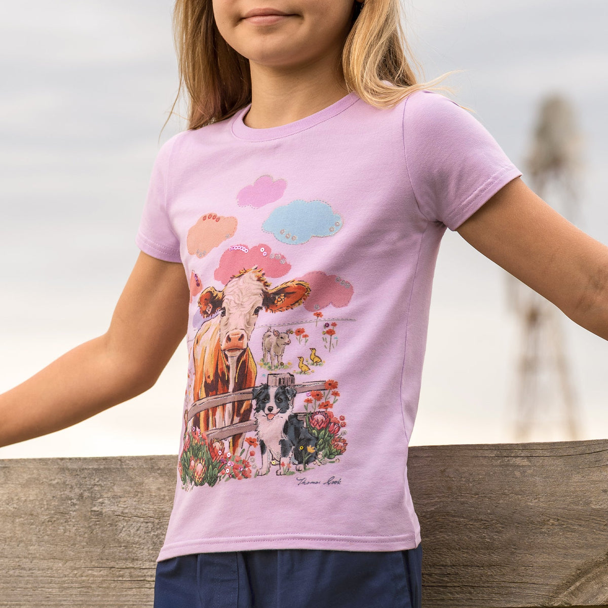 A young girl with blonde hair stands outdoors wearing a light purple short-sleeve t-shirt with a colorful cow graphic on the front, paired with navy blue pants.
