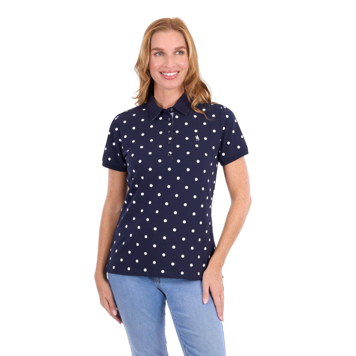 A woman modeling a short-sleeve polo shirt with a white polka dot pattern on a navy blue base, paired with light blue jeans, against a plain white background.